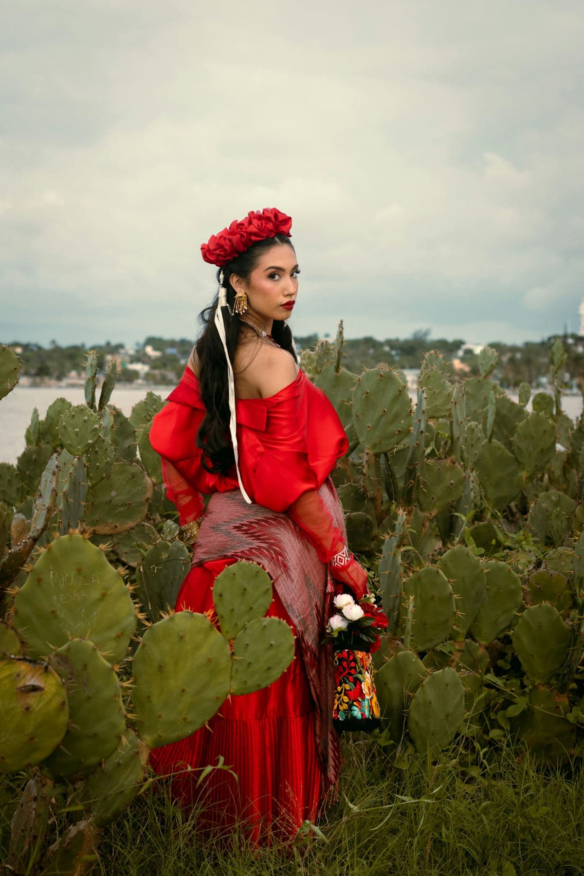 A woman in a red traditional Mexican dress with red flowers in her hair stands surrounded by tall green nopal cactus pads, holding a small bouquet